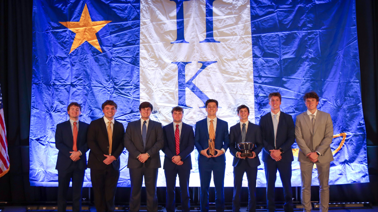 A group of nine people in formal suits stands on a stage in front of a large blue and white banner with a gold star with two individuals holding trophies.