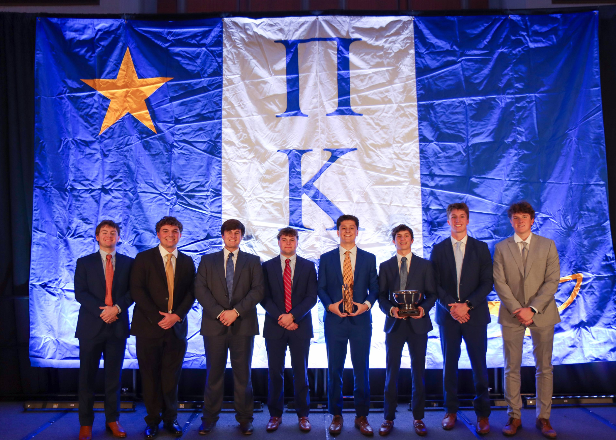 A group of nine people in formal suits stands on a stage in front of a large blue and white banner with a gold star with two individuals holding trophies.