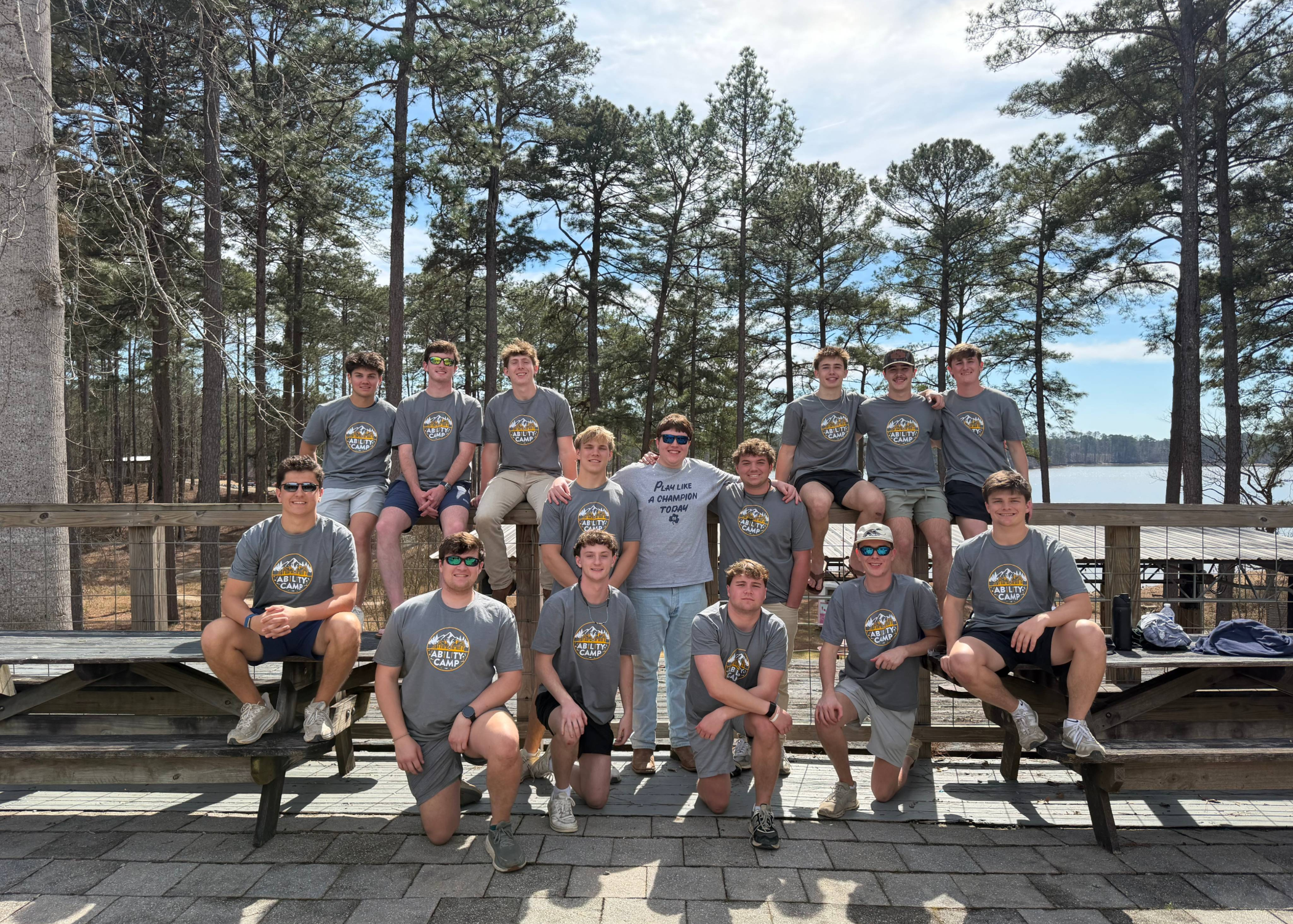 A group of about sixteen people in matching gray “Ability Camp” T-shirts pose on and around wooden benches on a deck overlooking trees and a lake under a partly cloudy sky.
