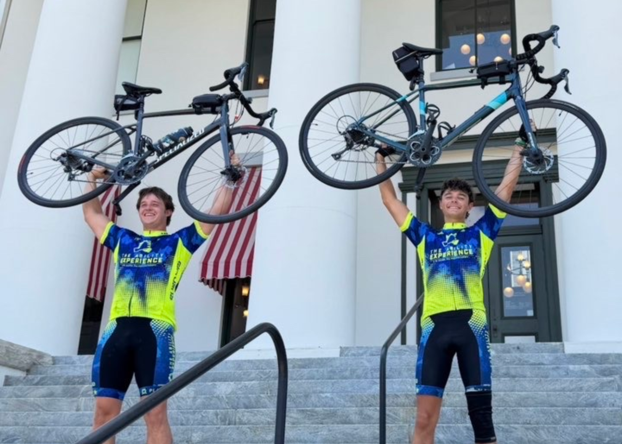 Two people wearing matching cycling kits stand on steps in front of a white-columned building, each holding a bicycle overhead.