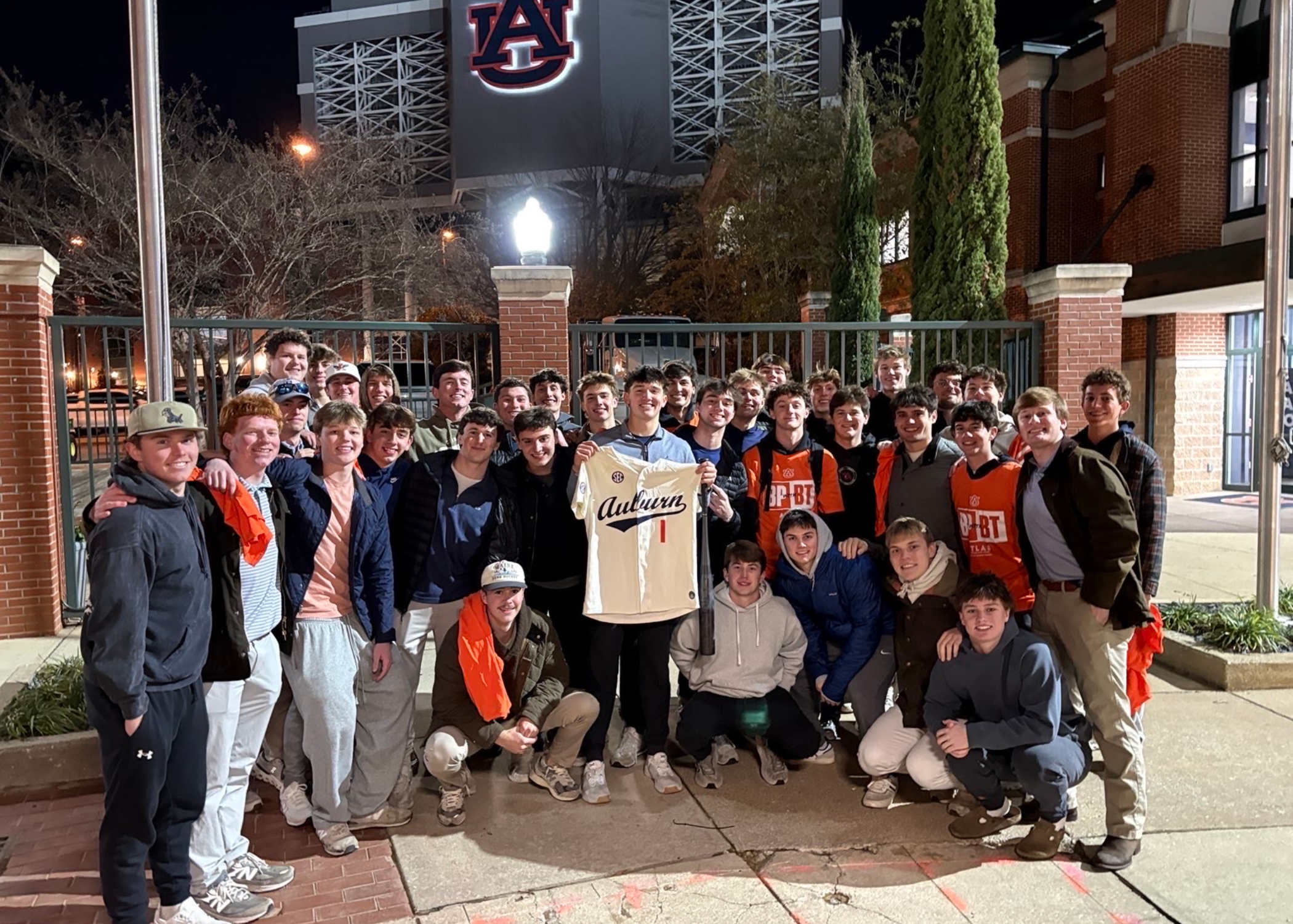 A large group of people stands outdoors at night in front of an Auburn University building, with one person holding up an Auburn jersey while others gather closely around.