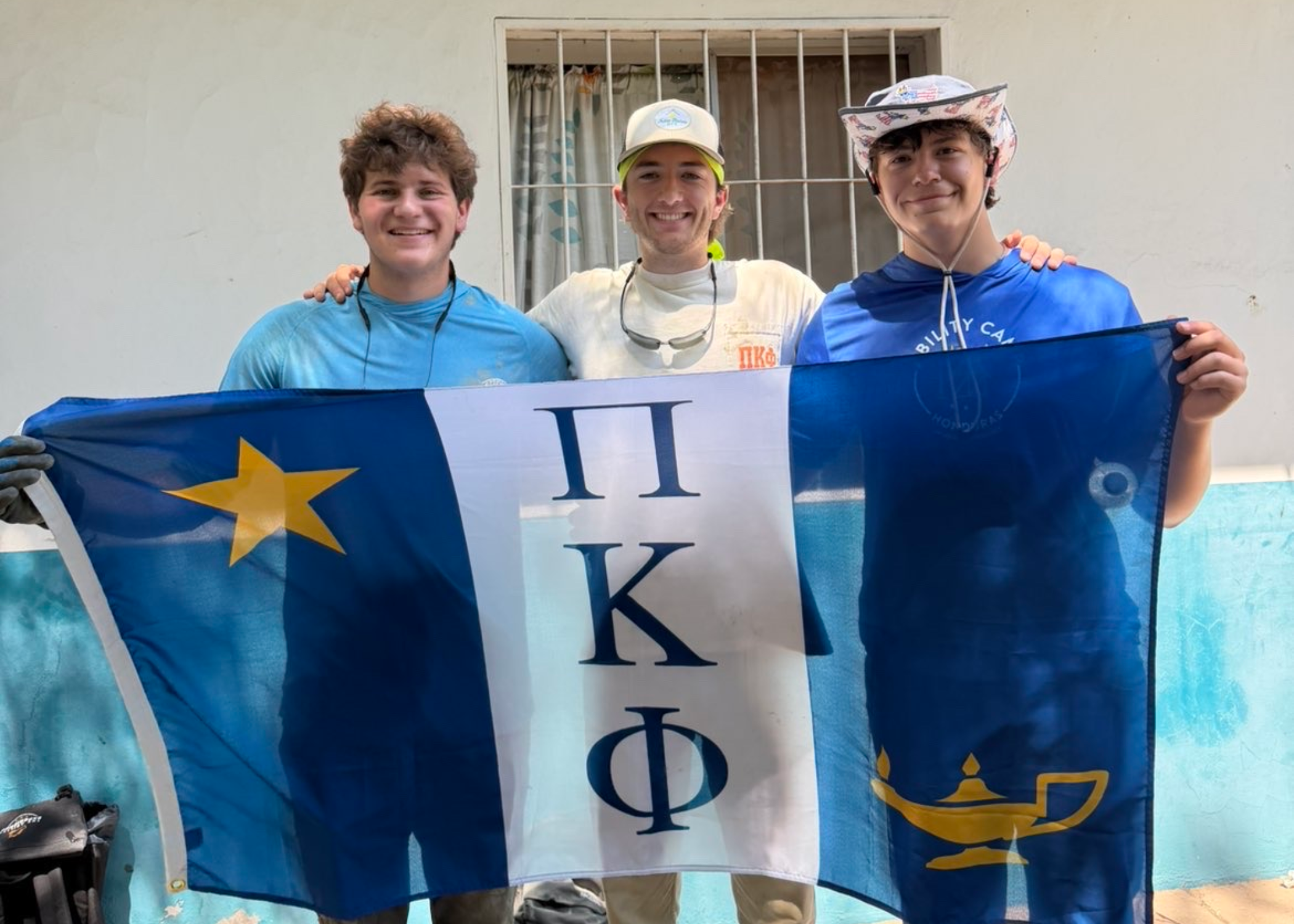Three people stand in front of a building holding a blue, white, and gold flag with Greek letters while posing side by side.