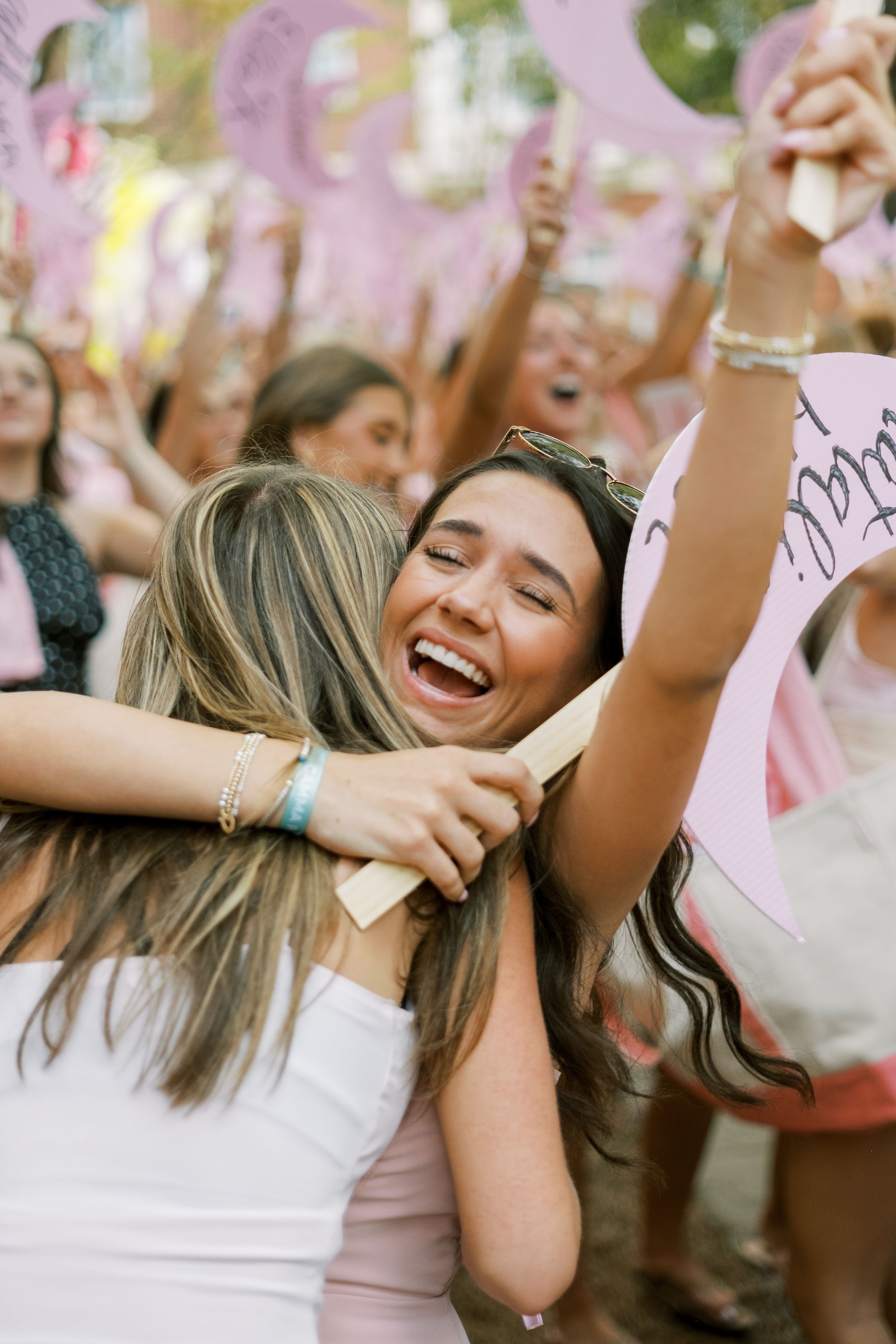 Two students embrace during a sorority bid day celebration, surrounded by a crowd holding pink signs and cheering outdoors.