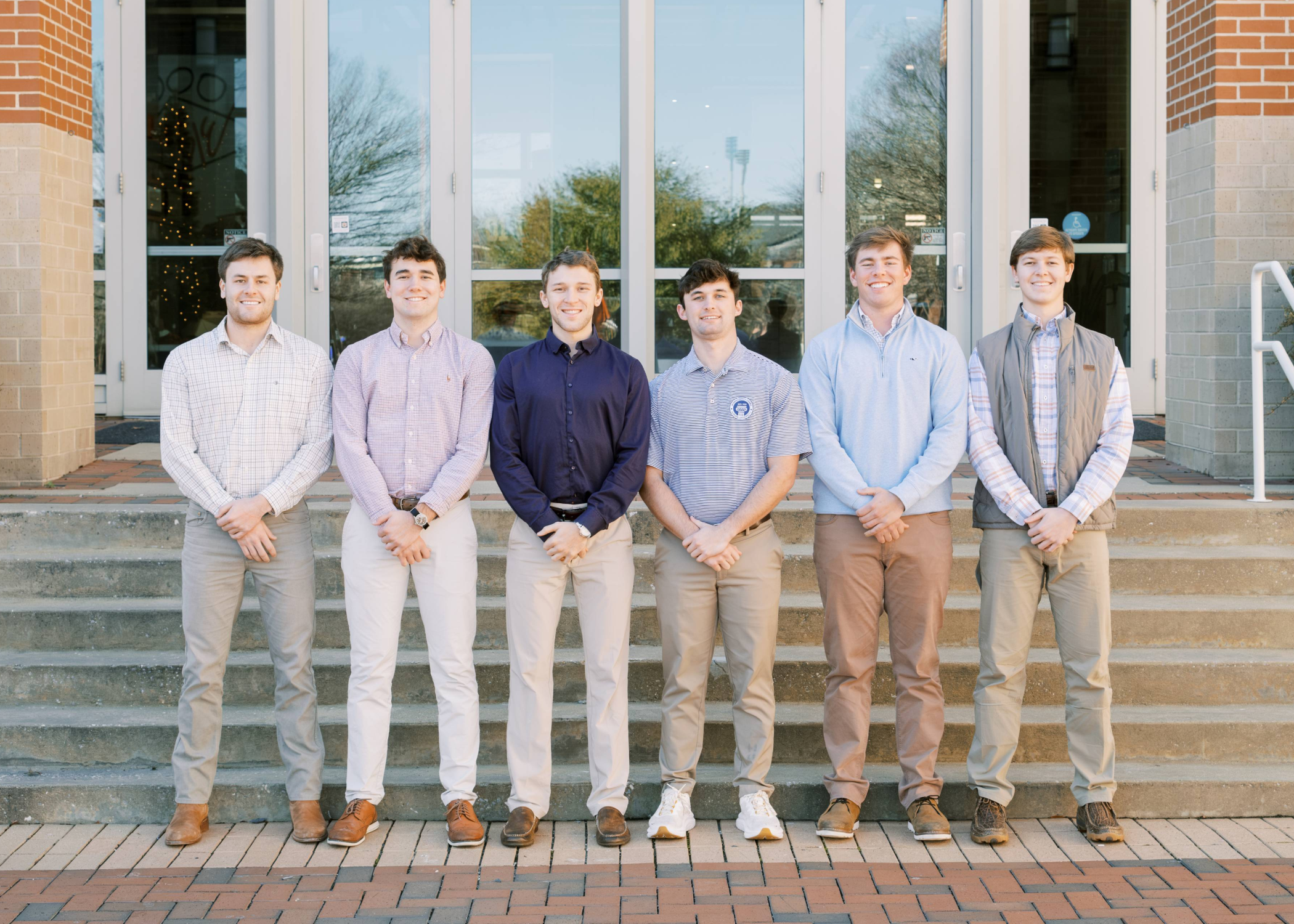 Seven people stand side by side on outdoor steps in front of a building entrance, dressed in casual business attire and facing the camera.