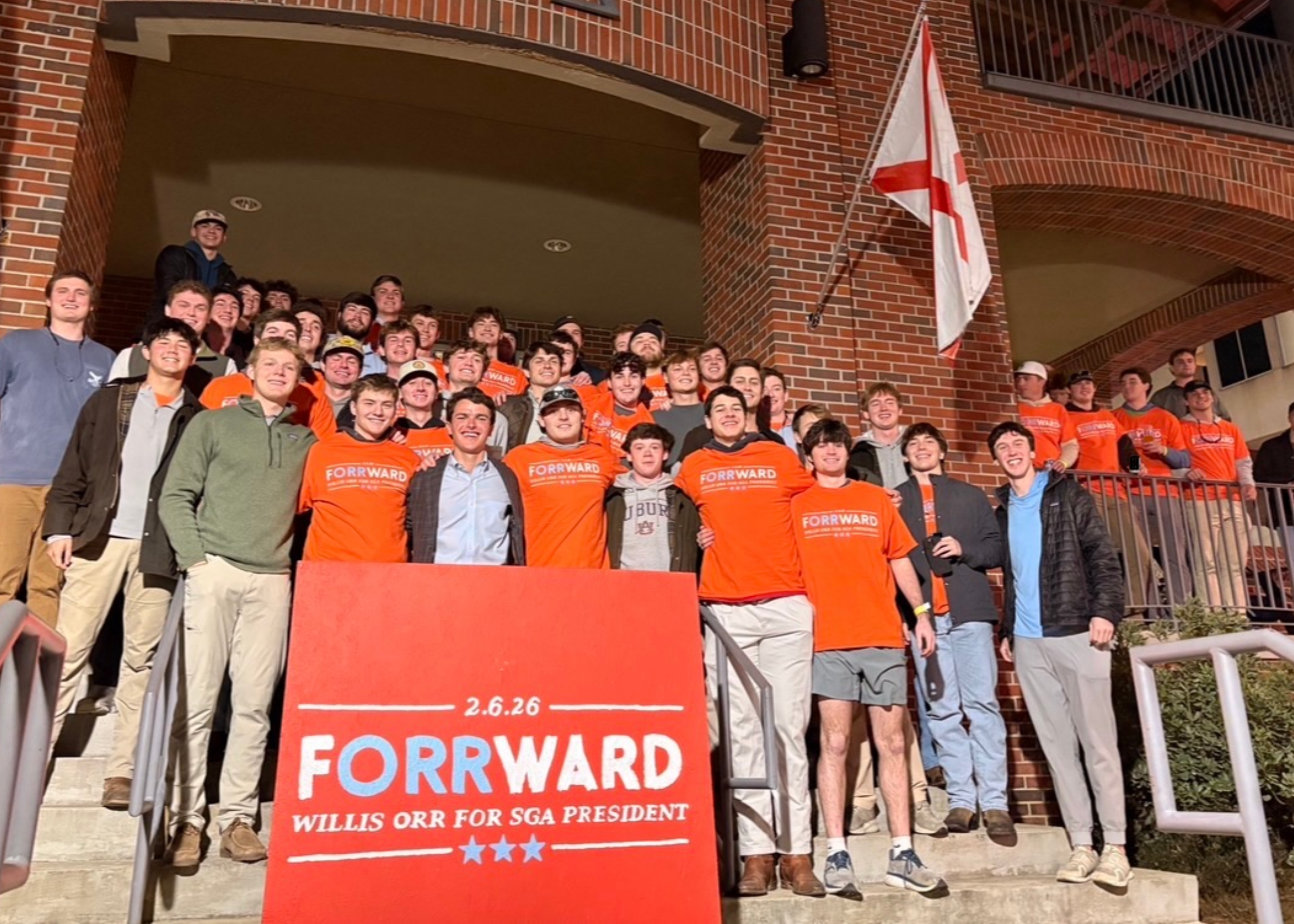 A large group of people stand together on brick steps outside a building, many wearing orange shirts, behind a red sign that reads “FORWARD Willis Orr for SGA President” .
