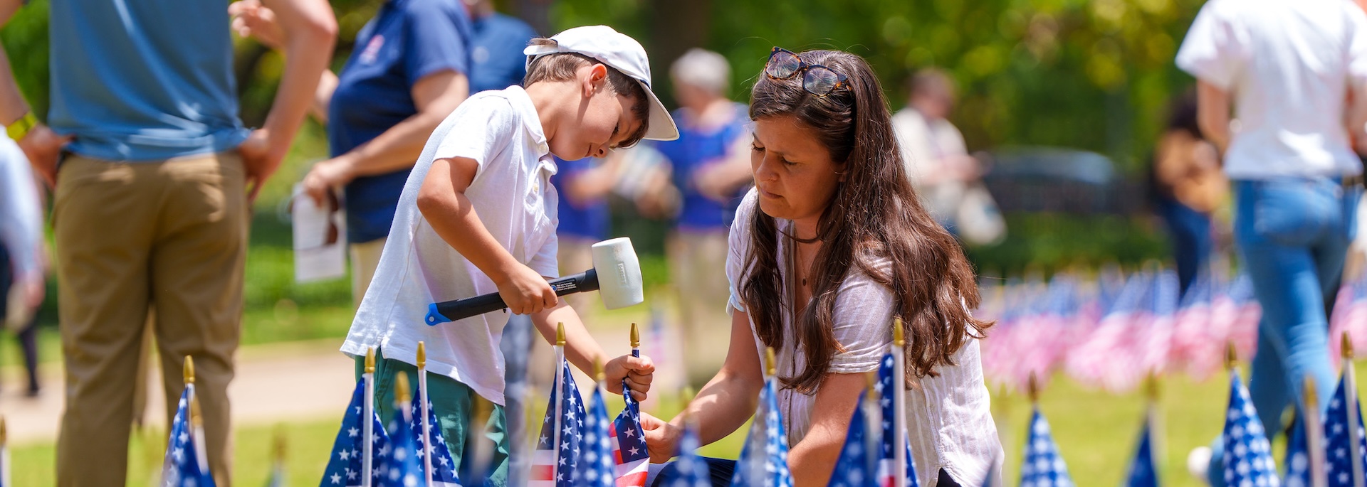 a mother holds a small flag as her son uses a mallet to place it in the ground