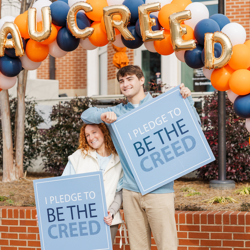 two students under an orange and blue balloon arch holding signs that say I pledge to be the creed