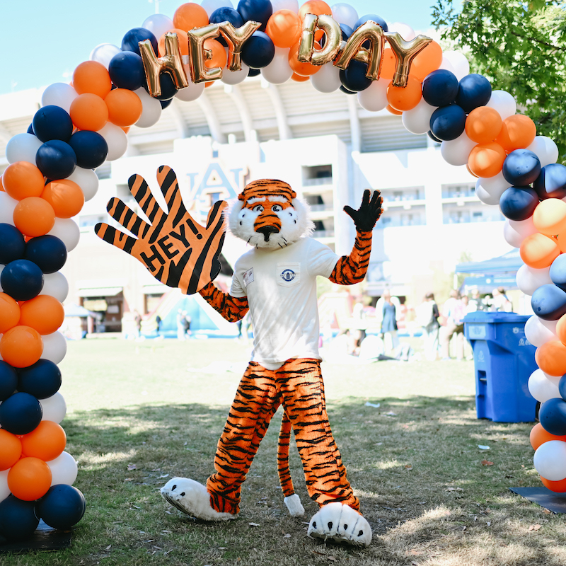 Aubie holding a giant foam hand with the word Hey on it under an orange and blue balloon arch
